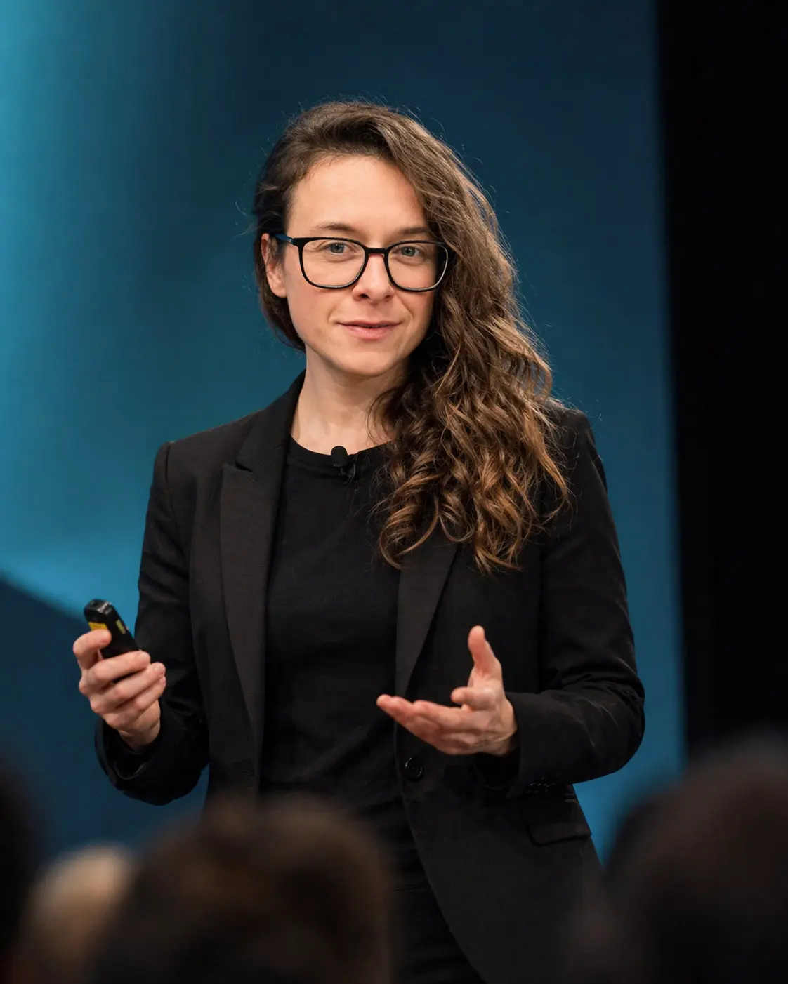 Laurence Paquette in a black blazer and glasses, holding a presentation clicker as she gives a keynote on a blue-lit stage