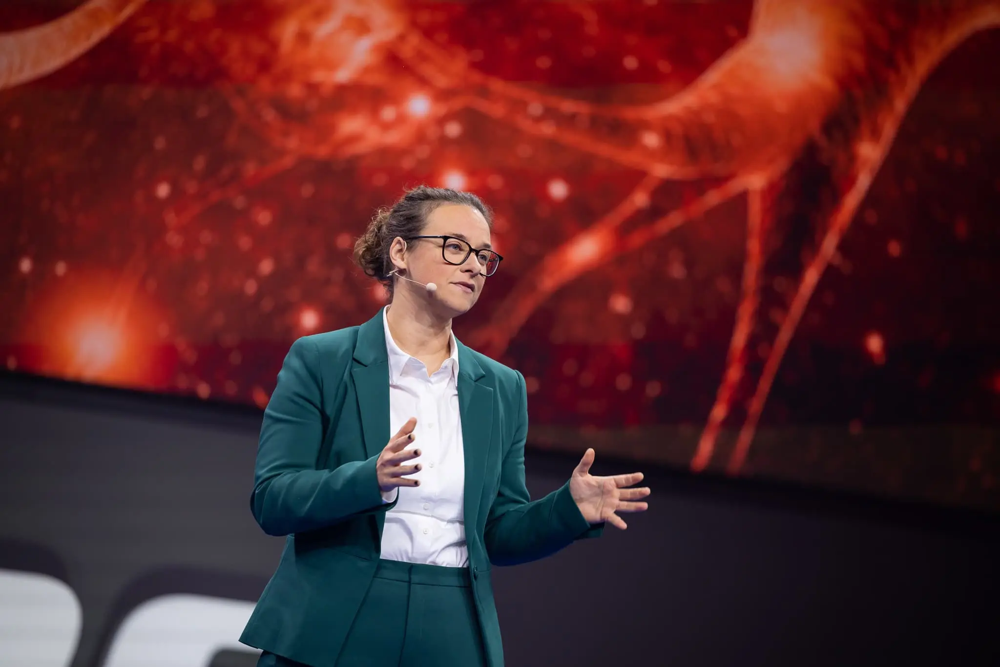 Laurence Paquette in a teal suit and headset microphone, gesturing as she speaks in front of a large red screen