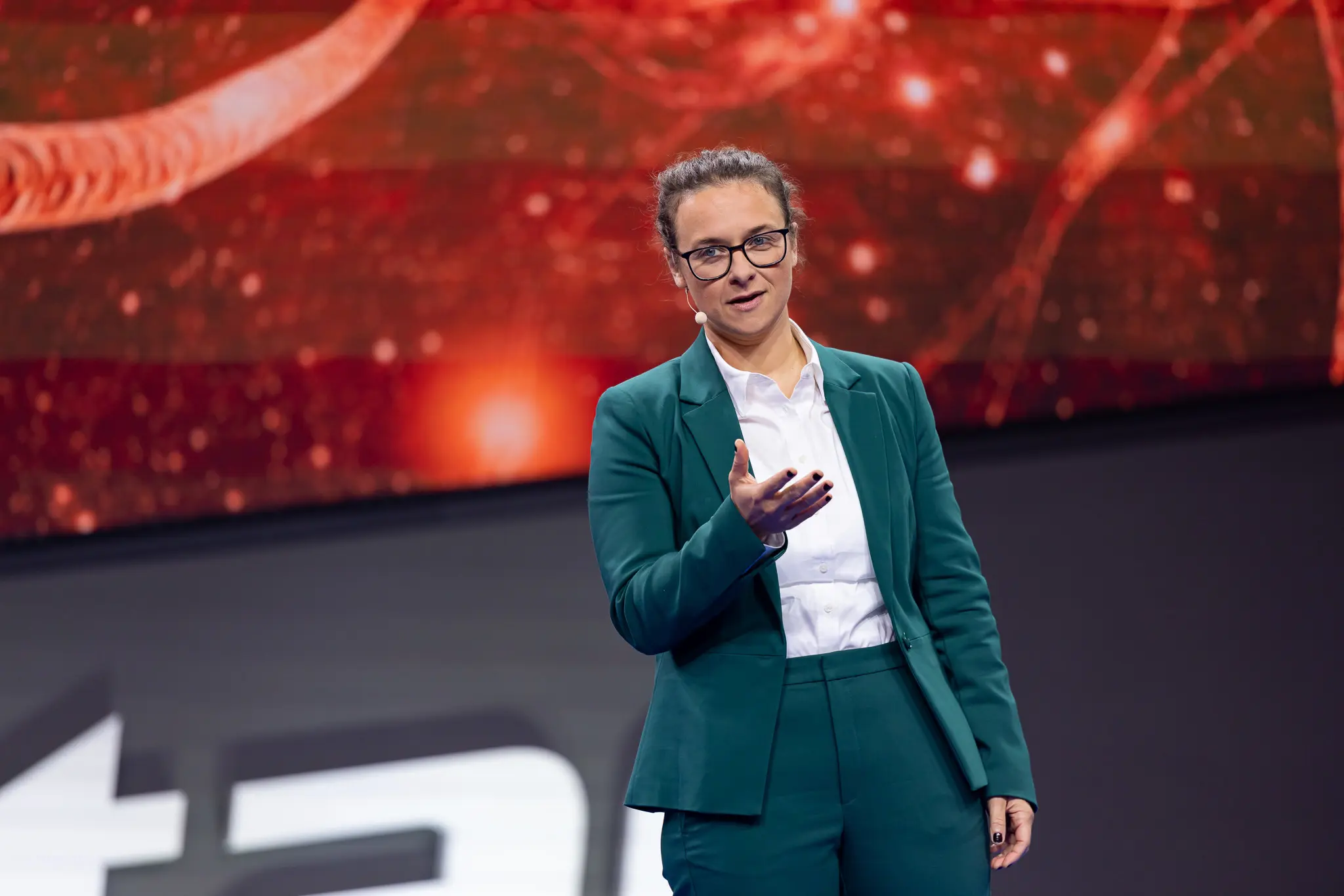 Laurence Paquette giving a talk on a red-lit stage in a teal suit, holding a microphone
